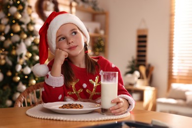 Cute little girl in Santa hat with delicious Christmas cookie and milk at home, space for text