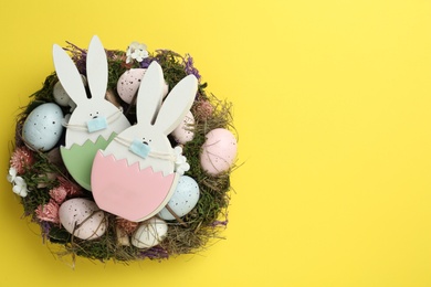 Wooden bunnies with protective masks, painted eggs, Easter wreath and space for text on yellow background, top view. Holiday during COVID-19 quarantine