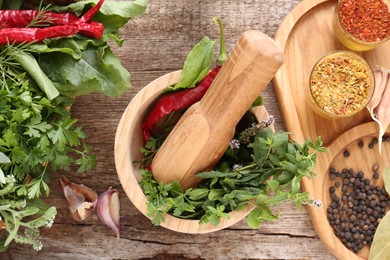 Mortar with pestle, fresh green herbs and different spices on wooden table, flat lay