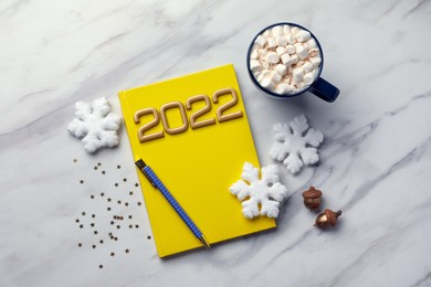 Stylish planner, cup of tasty hot drink and Christmas decor on white marble background, flat lay. 2022 New Year aims