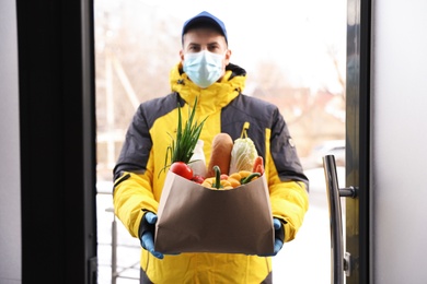 Courier in medical mask holding paper bag with groceries at doorway. Delivery service during quarantine due to Covid-19 outbreak