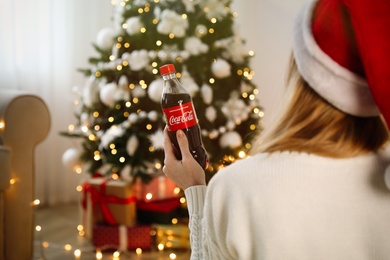 MYKOLAIV, UKRAINE - January 01, 2021: Woman in Santa hat with bottle of Coca-Cola against blurred Christmas tree at home, back view