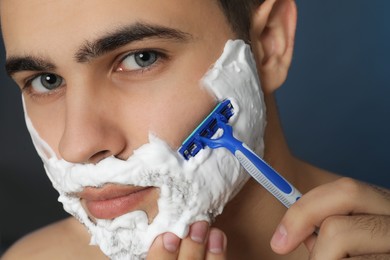 Handsome young man shaving with razor on blue background, closeup