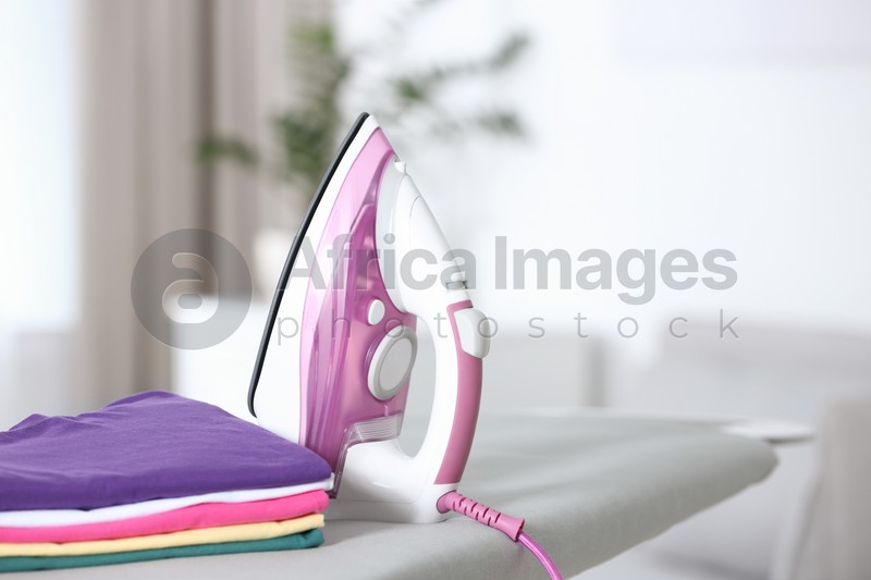 Modern electric iron and clean folded clothes on board in room. Space for text