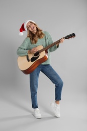 Young woman in Santa hat playing acoustic guitar on light grey background. Christmas music