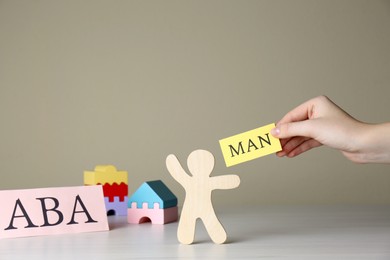 Photo of Woman holding paper with word Man near human figure and abbreviation ABA (Applied behavior analysis) at white wooden table, closeup