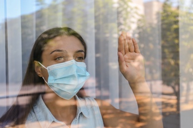 Stressed woman in protective mask looking out of window, view through glass. Self-isolation during COVID-19 pandemic