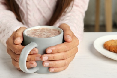 Woman holding cup of delicious cocoa drink at white table, closeup