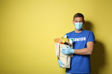 Male volunteer in protective mask and gloves with products on yellow background, space for text. Aid during coronavirus quarantine