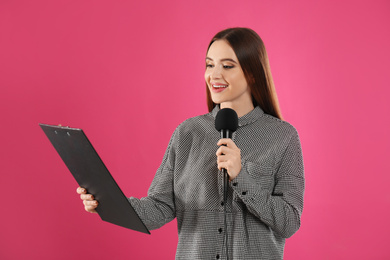 Young female journalist with microphone and clipboard on pink background