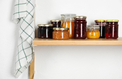 Jars with different homemade preserves on wooden shelf near white wall