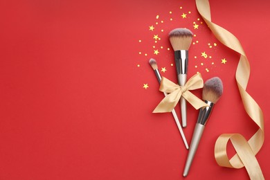 Different makeup brushes, ribbons and shiny confetti on red background, flat lay. Space for text