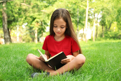 Cute little girl reading book on green grass in park