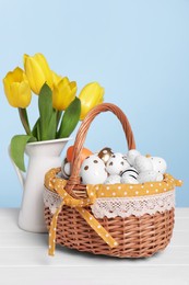 Photo of Wicker basket with festively decorated Easter eggs and beautiful tulips on white wooden table against light blue background