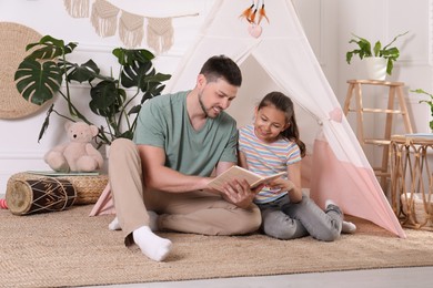 Father and daughter reading book near toy wigwam at home