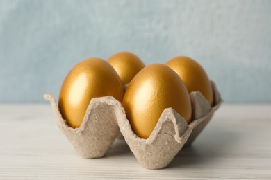 Photo of Carton with golden eggs on white wooden table, closeup