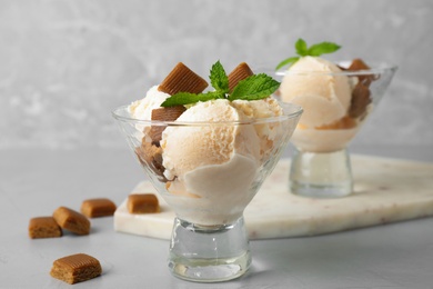Glass dish of delicious ice cream with caramel candies and mint on light grey table
