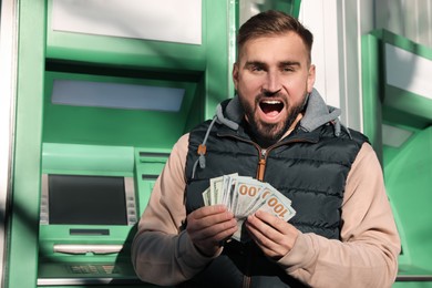 Photo of Excited young man with money near cash machine outdoors
