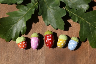 Colorful painted acorns with polka dot pattern and green oak leaves on wooden table, flat lay