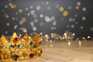 Photo of Beautiful golden crown with gems and fairy lights on wooden table, space for text. Fantasy item