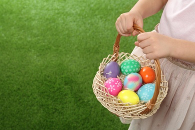 Little girl with basket full of Easter eggs on green grass, closeup. Space for text