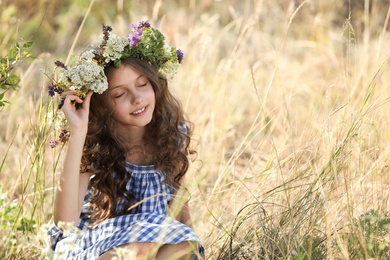 Cute little girl wearing wreath made of beautiful flowers in field on sunny day