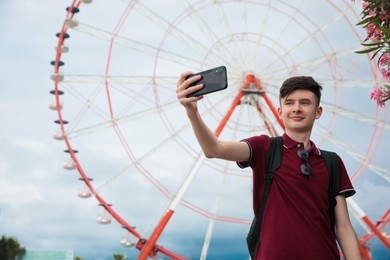 Teenage boy taking selfie near Ferris wheel outdoors. Space for text