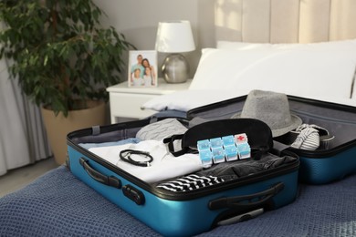 Open suitcase with packed clothes, accessories and pill box on bed indoors