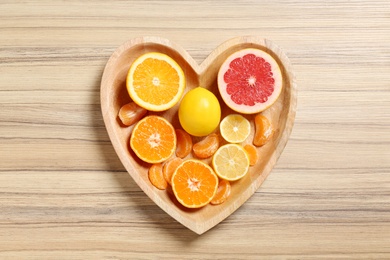Photo of Many fresh citrus fruits in heart shaped box on wooden background, top view
