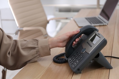 Woman using desktop telephone at wooden table in office, closeup. Hotline service