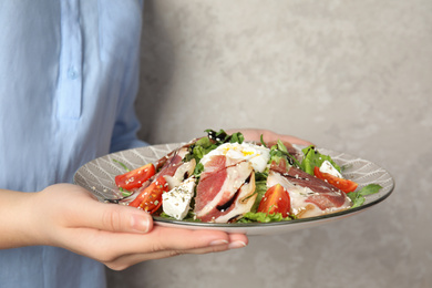 Woman holding plate with delicious prosciutto salad on grey background, closeup
