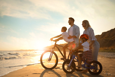 Happy parents teaching children to ride bicycles on sandy beach near sea at sunset