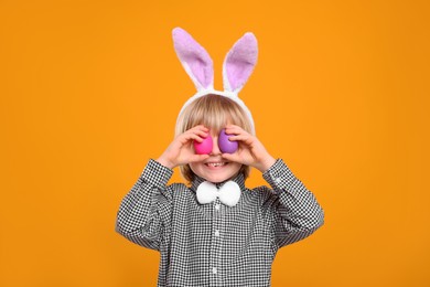 Photo of Happy boy in bunny ears headband holding painted Easter eggs near his eyes on orange background