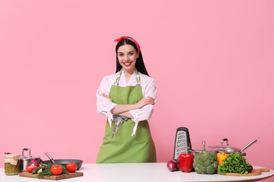 Young housewife at white table with utensils and products on pink background