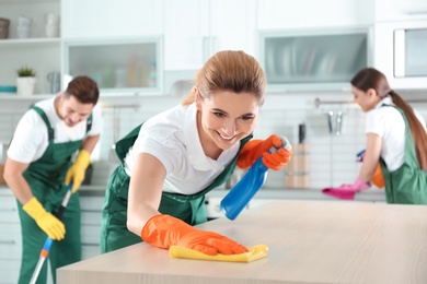 Woman using rag and sprayer for cleaning table with colleagues in kitchen