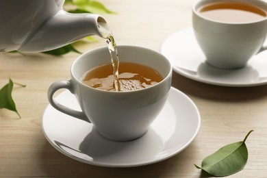 Pouring green tea into white cup with saucer and leaves on wooden table, closeup