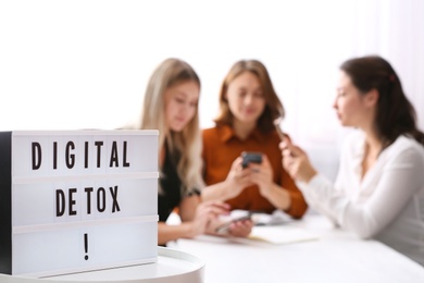 Women using smartphones at table in office, focus on lightbox with phrase DIGITAL DETOX