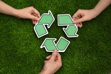 People holding paper recycling symbol above green grass, top view
