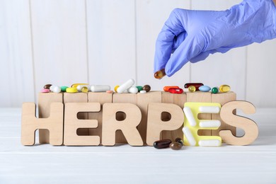 Woman in glove putting pill near word Herpes on white wooden table, closeup