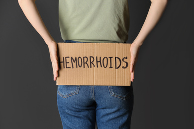 Photo of Woman holding carton sign with word HEMORRHOIDS on black background, closeup