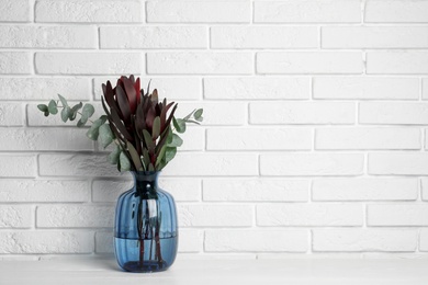 Bouquet of beautiful protea flowers and eucalyptus branches in glass vase on white wooden table near brick wall. Space for text
