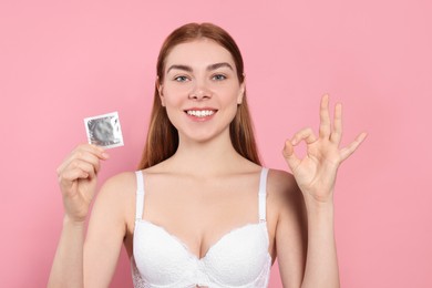 Photo of Woman in bra with condom showing ok gesture on pink background. Safe sex