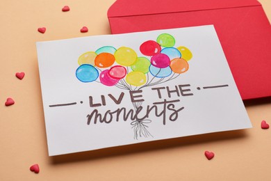 Card with phrase Live The Moments and red envelope on beige background, closeup