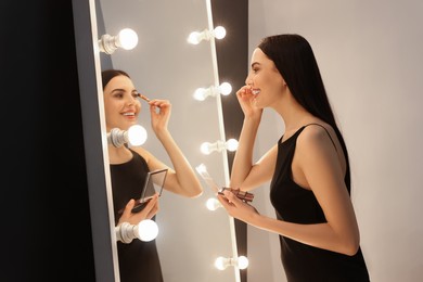 Photo of Beautiful young woman in elegant dress applying eyeshadow with brush near mirror indoors