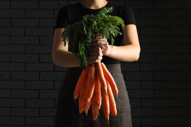 Photo of Woman holding fresh ripe juicy carrots against dark brick wall, closeup