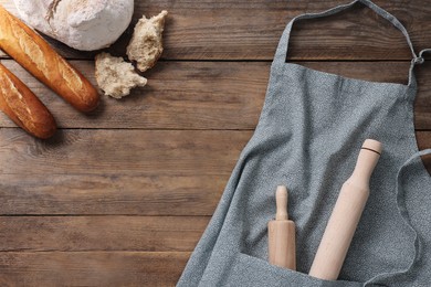 Clean kitchen apron with rolling pins and different types of bread on wooden table, flat lay. Space for text