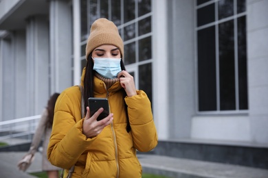Young woman in medical face mask with smartphone walking outdoors. Personal protection during COVID-19 pandemic