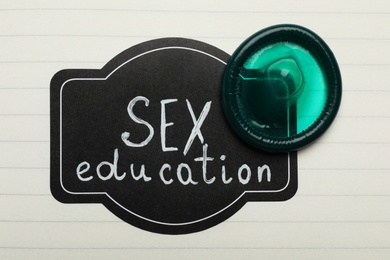 Green condom and tag with words SEX EDUCATION on paper sheet, top view