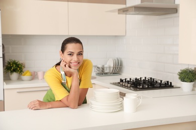 Beautiful young woman with clean dishes and cups at table in kitchen