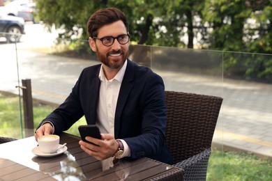 Handsome bearded man with cup of drink and smartphone at table in outdoor cafe